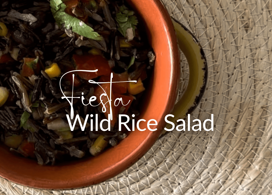 A close-up view of Fiesta Wild Rice Salad in a terracotta bowl. The salad features a mix of dark wild rice, black beans, yellow corn, and chopped green cilantro, creating a colorful contrast. The bowl sits on a textured beige woven mat, and the name 'Fiesta Wild Rice Salad' is elegantly scripted in white across the top of the image.